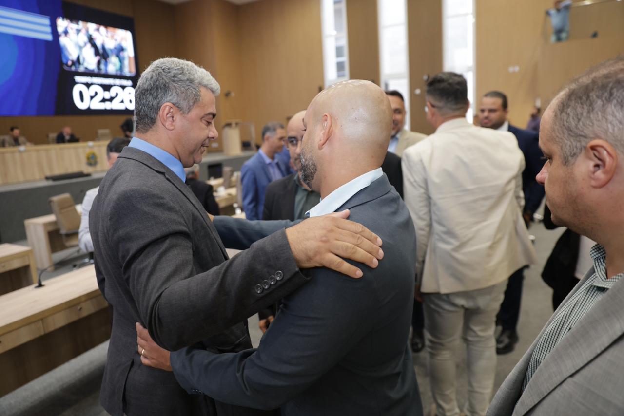 Homens conversam em conferência pública com painel ao fundo.
