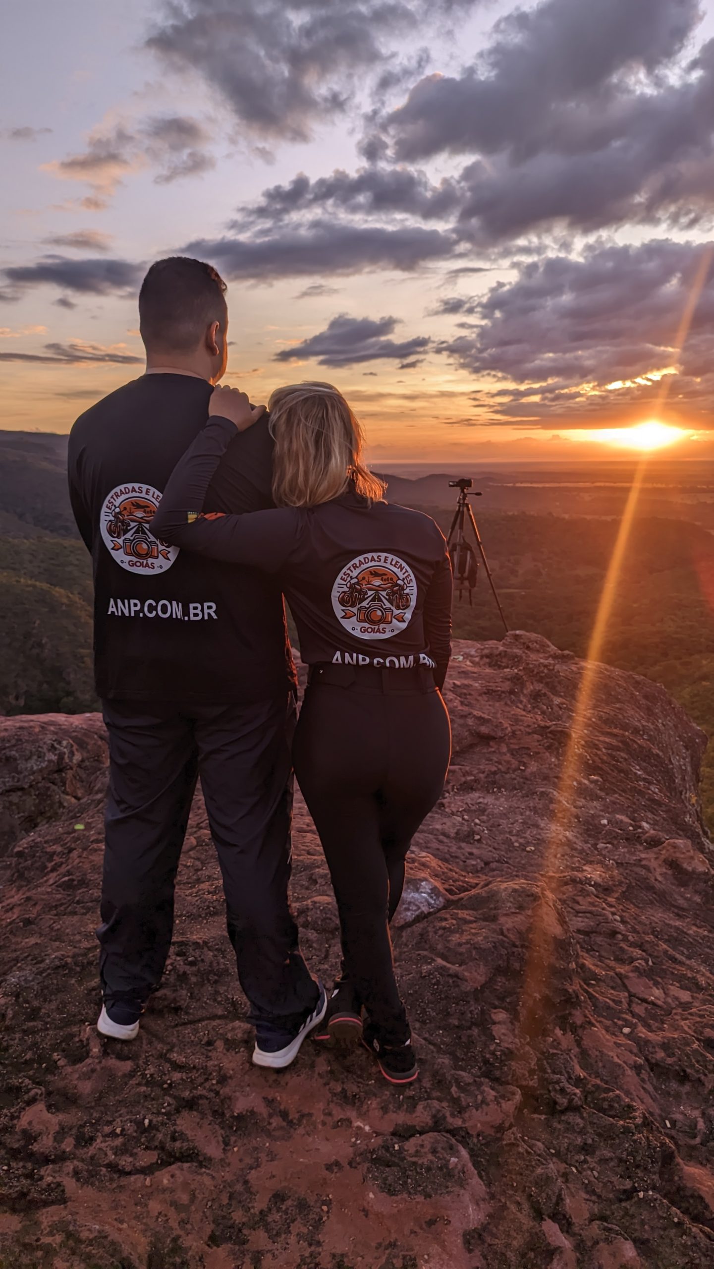 Casal observa pôr do sol em mirante rochoso.