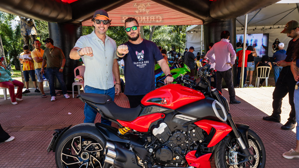 Dois homens posam com moto esportiva vermelha em evento.